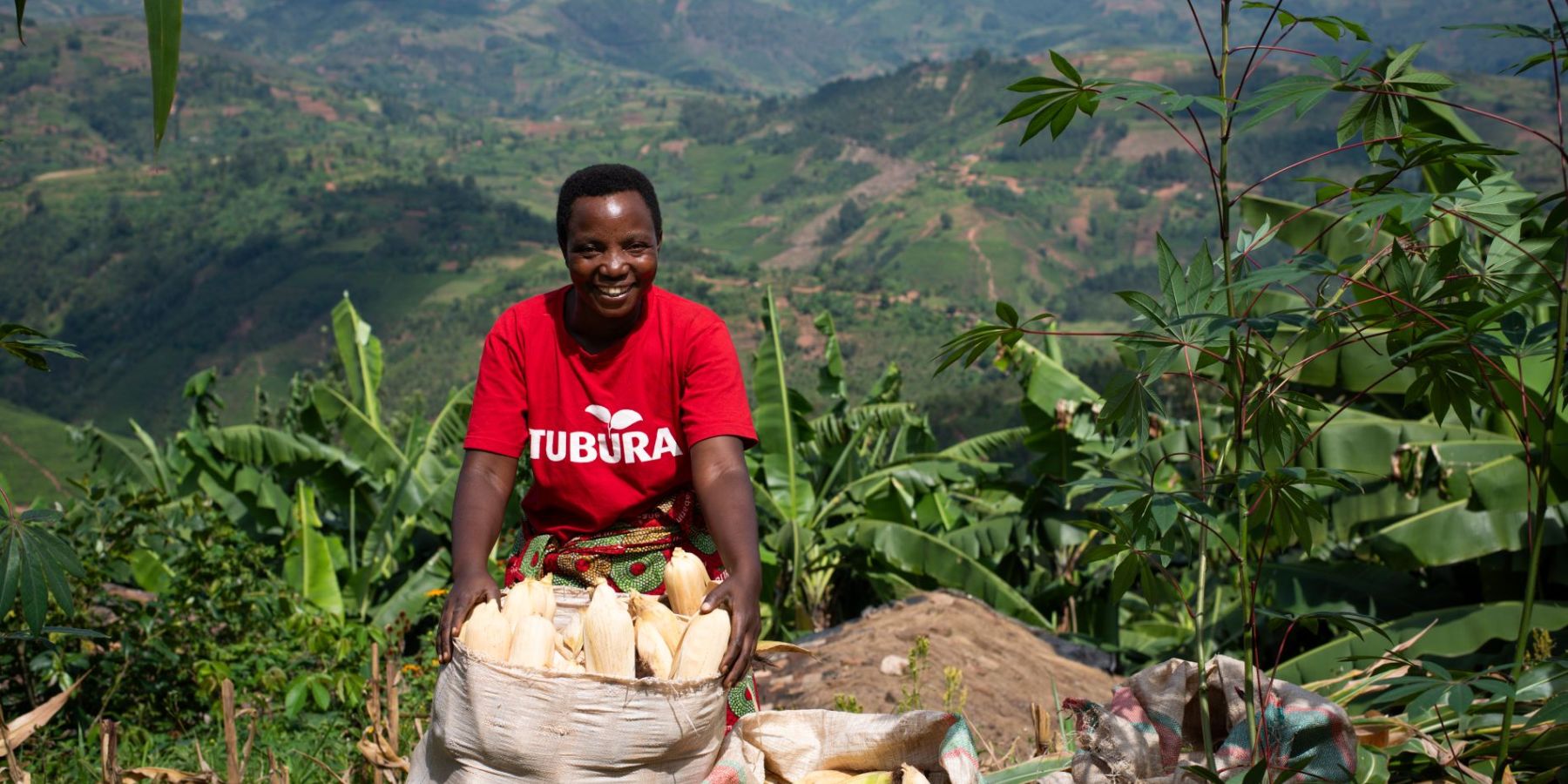 A smallholder farmer smiles in front of her maize harvest in sacks. Behind her are the green hills of Burundi.
