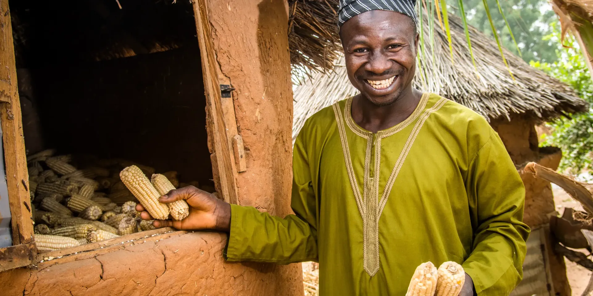 A smiling farmer holding maize in their hands and standing next to their harvest.
