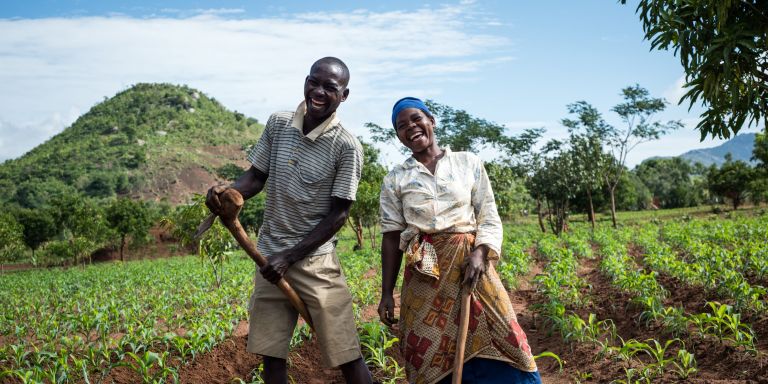 A husband and wife stand smiling in their field in Malawi