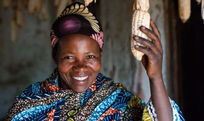 A picture of a smallholder farmer smiling and standing underneath maize drying from the roof. she is holding one cob of maize in her hand.