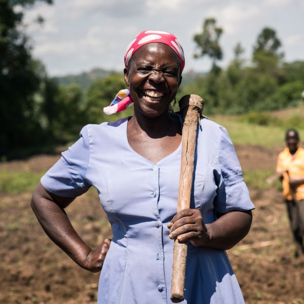 A farmers stands smiling in her field with her jembe slung over her shoulder