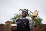 A farmer in Tanzania holding cobs of maize as she smiles at the camera