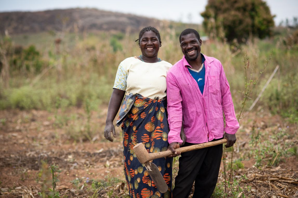 A couple stands in their field. One person holds a hoe.