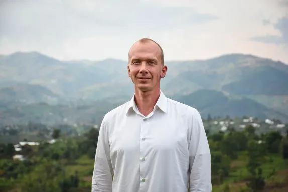 A man in a white shirt is standing with a hilly background